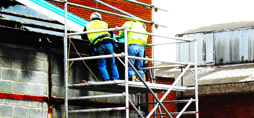 close up corner view of two brook scaffolding scaffolders erecting scaffold arround a factory built from breeze blocks with corrigated bright blue roofing